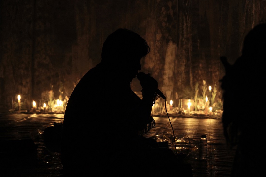 Silhouette of a seated singer in a dark ceremony space with faint candlelight behind