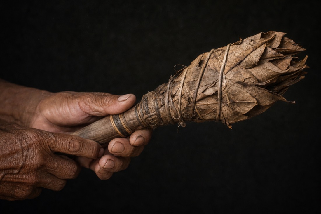 Weathered hands holding a leaf-bundle rattle against a plain dark background