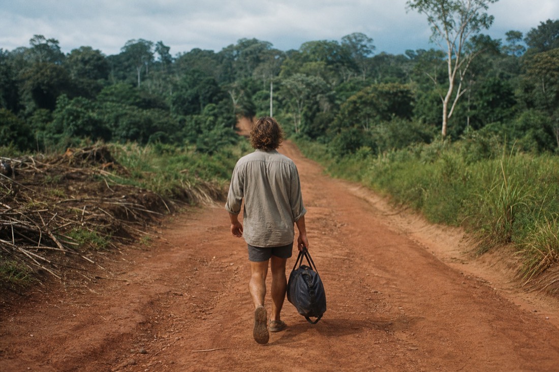 Backpacker walking down a dirt road toward a jungle treeline carrying a small duffel