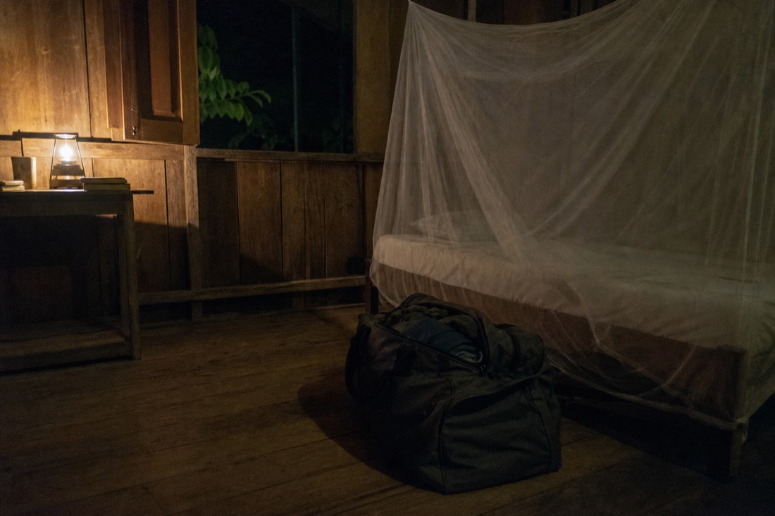 Packed duffel bag beside a single bed with mosquito net in low lamplight