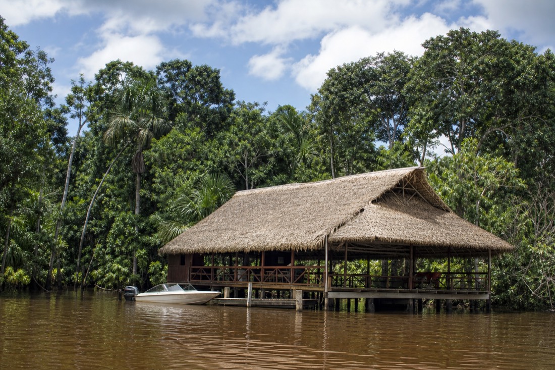Open-sided wooden retreat building beside a calm river with a moored speedboat, midday light, Amazon river near Iquitos