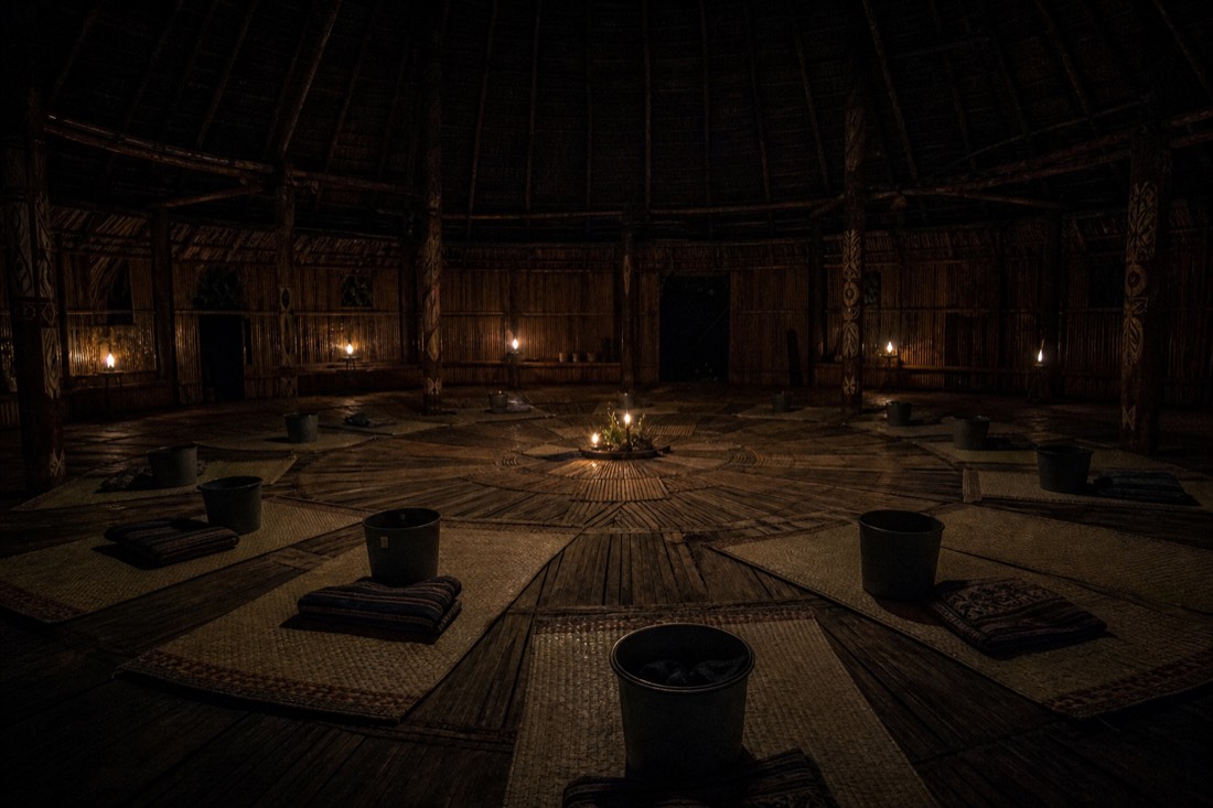 Simple ceremonial maloca interior at night with woven mats, small buckets and low candlelight, Iquitos region, no people