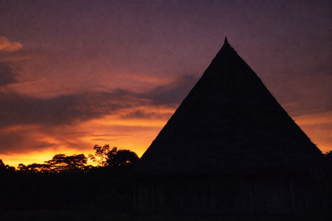 Last light fading over a thatched maloca roof silhouetted against a deep orange Amazon sky