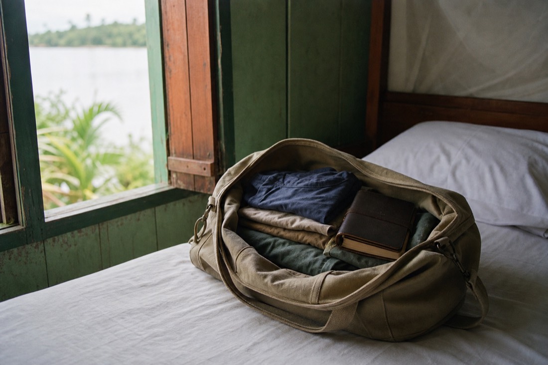 Open canvas duffel bag with folded clothes and a journal on a simple guesthouse bed