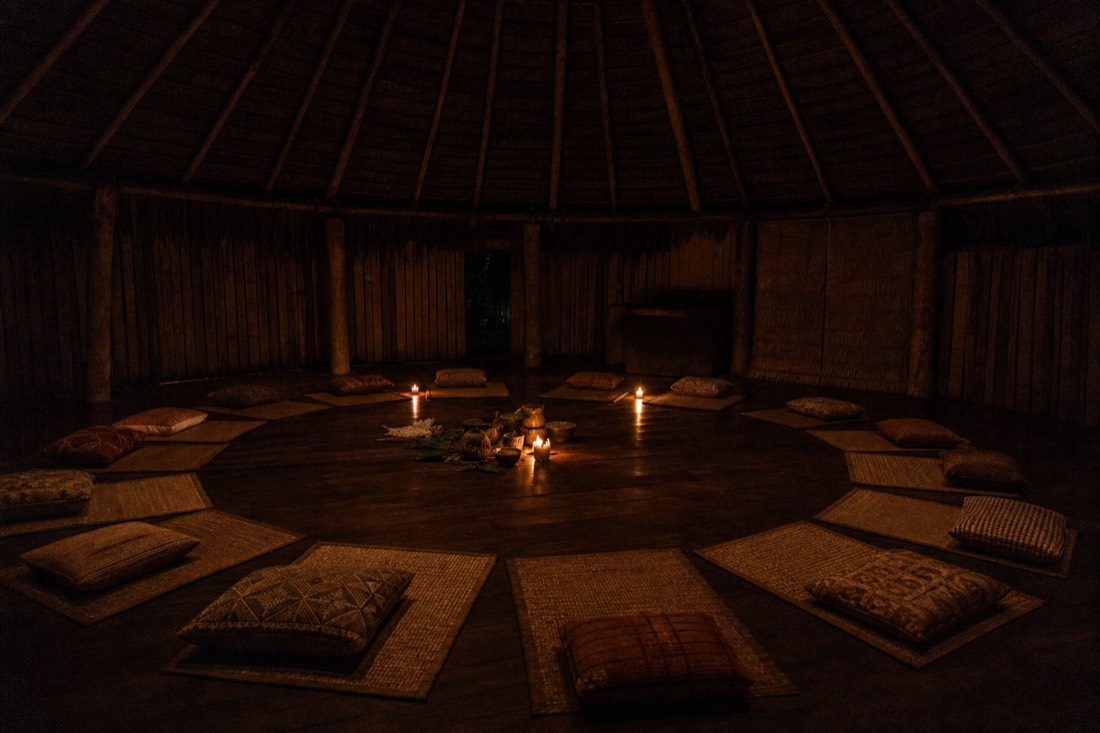 Candlelit ceremonial space inside a wooden maloca at night, empty mats and cushions arranged in a circle, Iquitos region