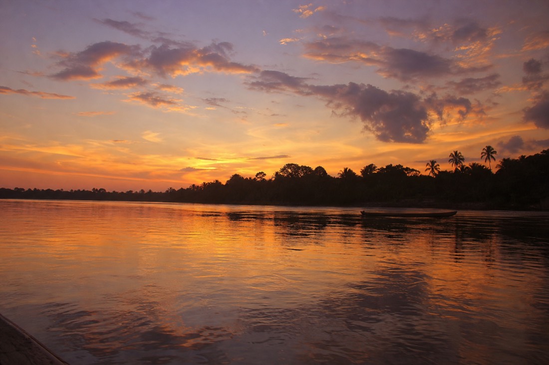 Sunset over the Amazon river with silhouetted palms and a distant wooden canoe, Loreto region, Peru